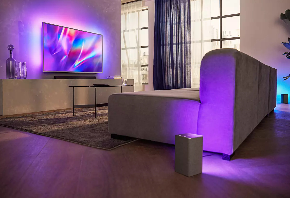 Απεικονίζεται το soundbar κάτω από τηλεόραση και το subwoofer δίπλα σε καναπέ.